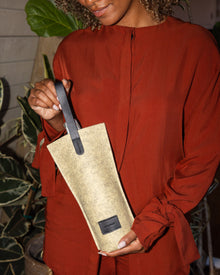 A person in a rust-colored outfit holds a graf lantz beige wine bag with elegant leather handles, standing in a room decorated with various green plants that enhance the stylish ambiance.