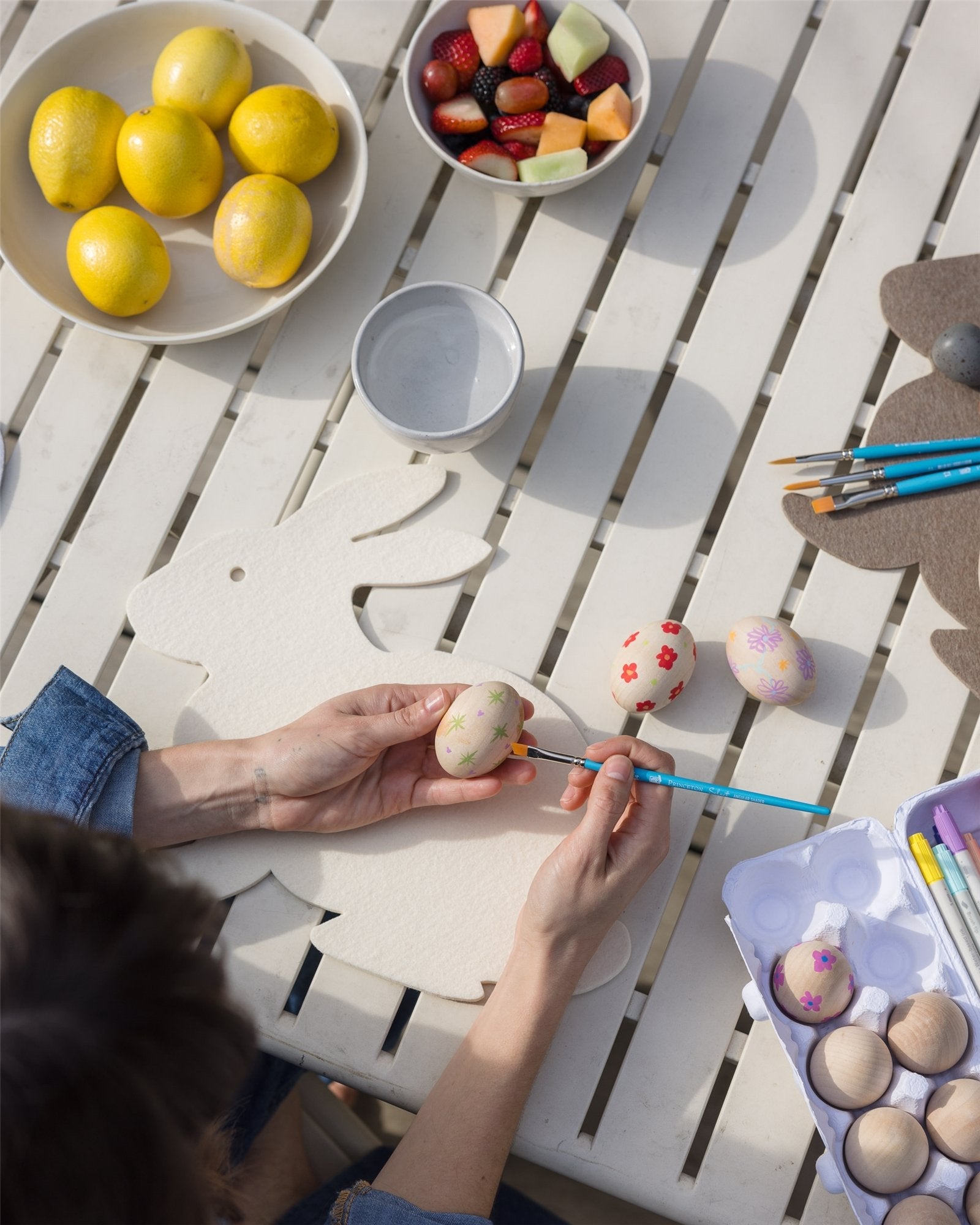 Someone is painting colorful patterns on eggs at a table featuring a heat-resistant Graf Lantz Merino Wool Bunny Trivet. Nearby are lemons, mixed fruit, and a paintbrush set, with a carton of decorated, season-exclusive eggs completing the scene.