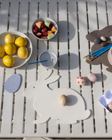 A Graf Lantz Merino Wool Bunny Trivet highlights a table of Easter craft supplies, including decorated eggs, paintbrushes, a palette, and bowls of lemons and mixed fruit, creating a festive and creative atmosphere.