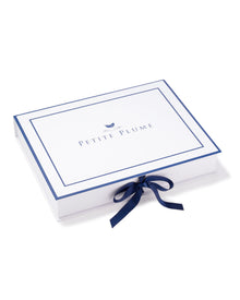 A rectangular box, featuring PETITE PLUME on the lid with a bird logo above, exudes luxury with its white and blue design and side bow. It encloses the Mens Silk Pajama Set in Black by Petite Plume, highlighting its machine-washable silk charm.