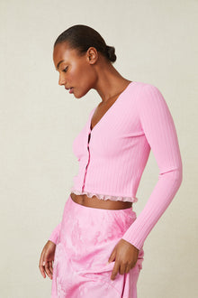 Side image of model wearing pink v-neck cardigan with ruffle detail at hem and button up closure.