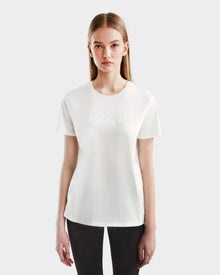 A young woman with straight, long blonde hair wears the Rudsak AMINA - 8524012 Cotton White women’s crewneck short sleeve t-shirt with a subtle front embossing, paired with black pants, standing against a plain white background.