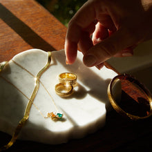 Flatlay Model Close-up of a woman's hand reaching toward gold rings and a necklace resting on a white marble surface.