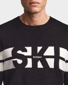 A person wears the OMAR - 6523507 Black men’s knit crewneck sweater by Rudsak, featuring bold white SK letters and the brand name across the chest. The image shows only the lower face, neck, and upper chest.