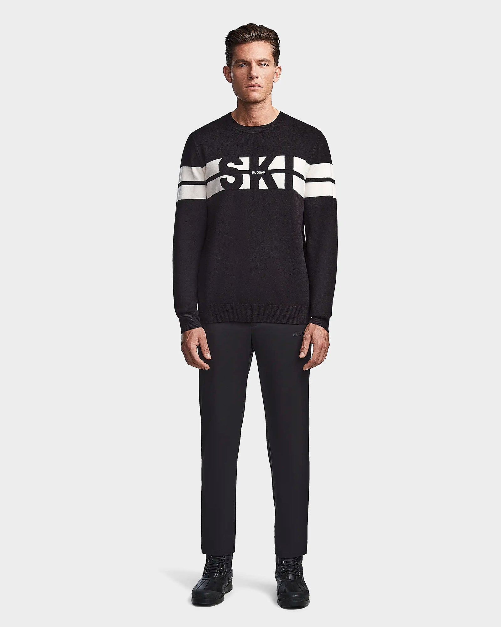 A man faces forward wearing the Rudsak OMAR - 6523507 Black mens knit crewneck sweater with white sleeve stripes and SKI on the chest, paired with black pants and shoes, set against a plain light background.