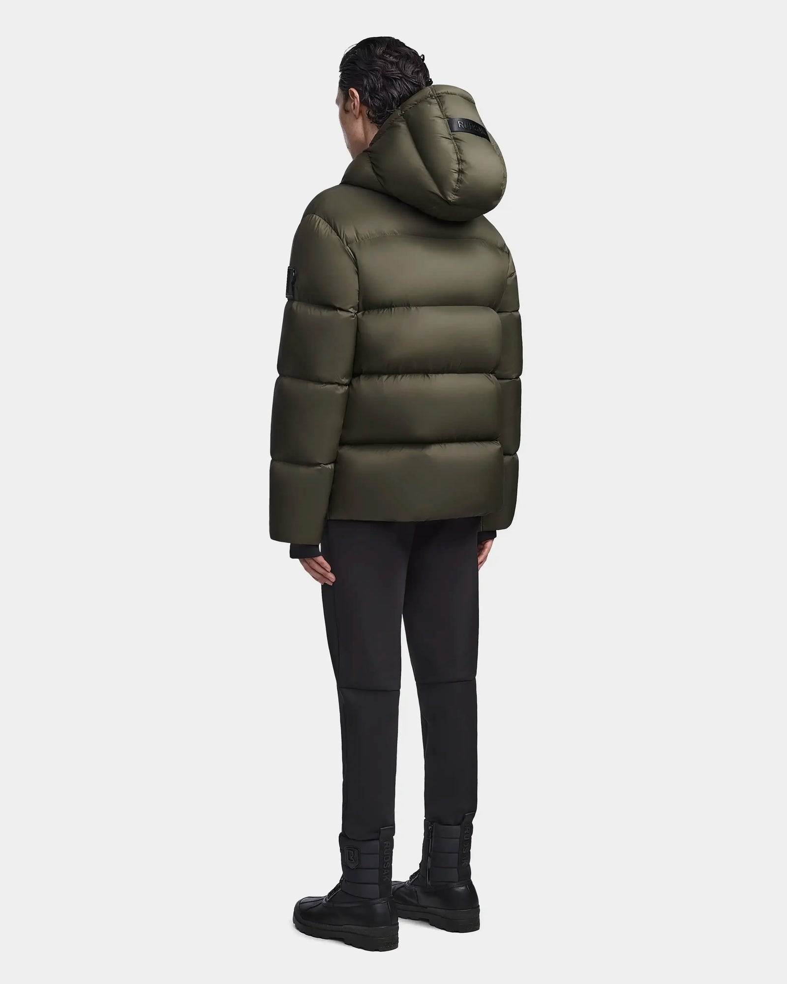 A person stands facing away, wearing the Rudsak DANIEL M - M123510 Light Olive mens hooded down puffer with black pants and boots against a plain light background.