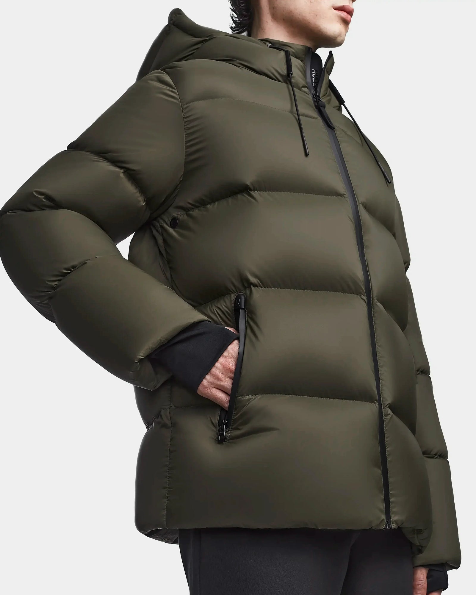 A person wears the Rudsak DANIEL M - M123510 Light Olive men’s hooded down puffer with a black zipper and zippered pockets. Its quilted, padded design offers warmth and weather resistance, shown from shoulders to mid-thigh.