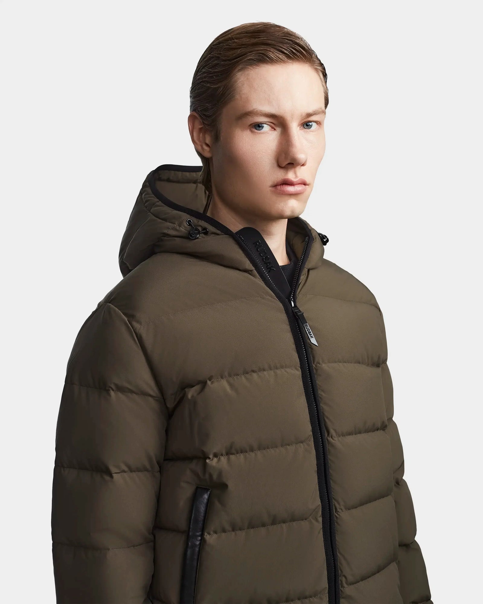 A young person with light skin and straight, light brown hair pulled back wears the Rudsak DEMIAN M - M123507 Light Olive hooded down puffer, made from sustainable recycled fabric with a black zipper, against a plain light background.