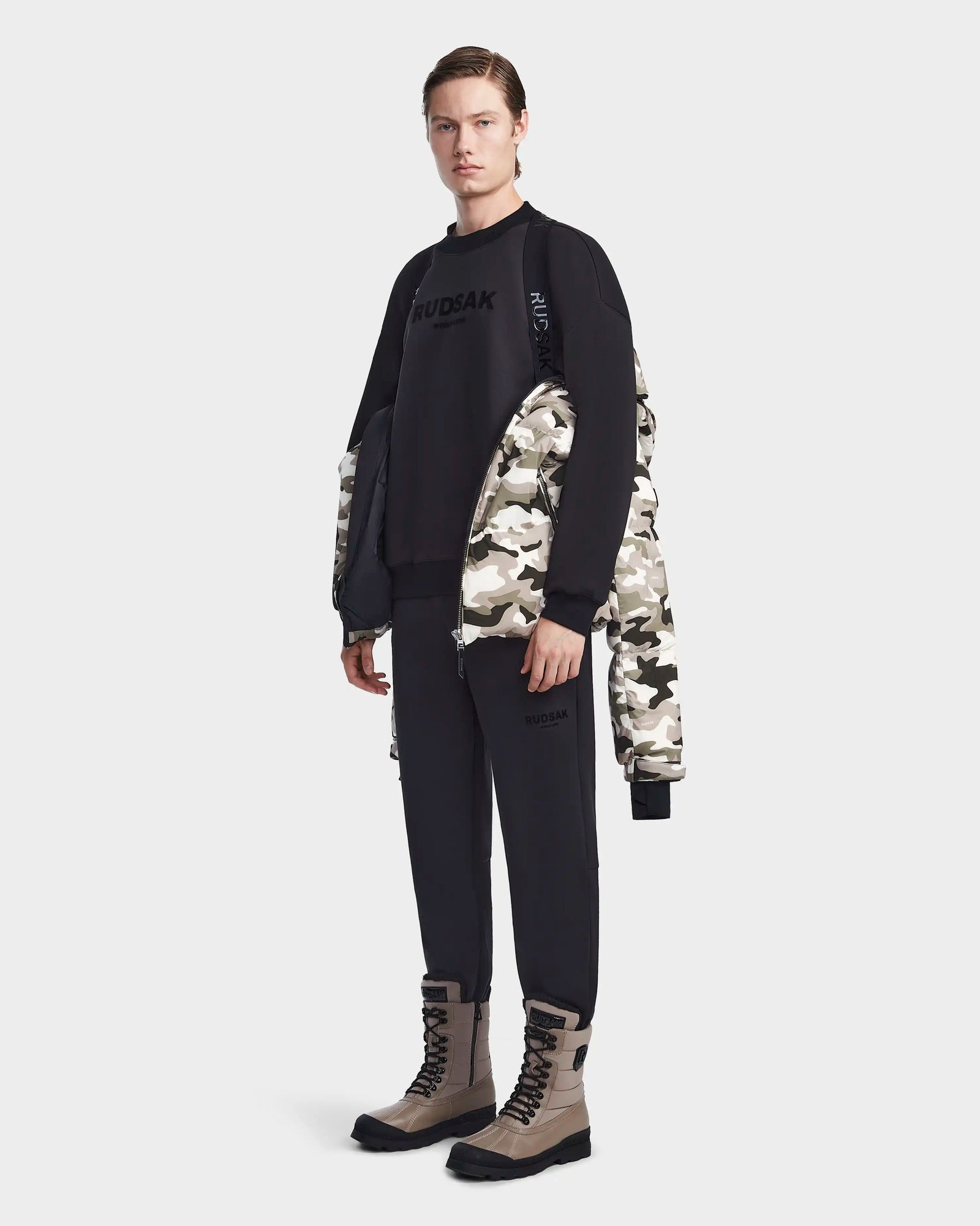 A person stands against a plain light background wearing the Rudsak VINCENT M - 6123510 Camouflage water-repellent jacket with lightweight insulation draped over their shoulders, paired with a black sweatshirt, pants, and beige and black boots.