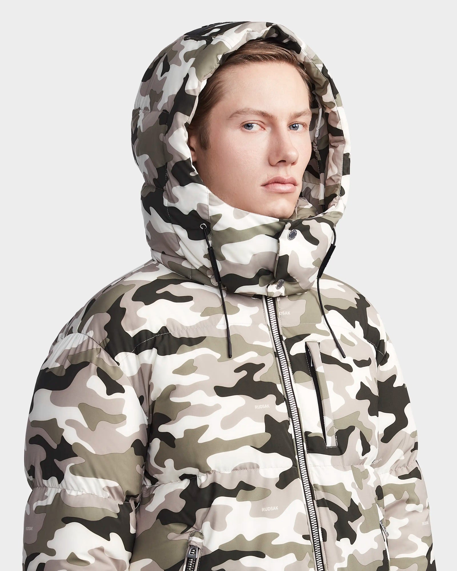 A person wearing the Rudsak VINCENT M - 6123510 Camouflage men's down puffer with lightweight insulation and the hood up, looking slightly to the side against a plain light background.