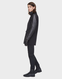 A person with long brown hair stands in profile against a plain light background, wearing the Rudsak BEAU - M121504 BLACK men’s coat with leather sleeves, black gloves, fitted black pants, and black boots.