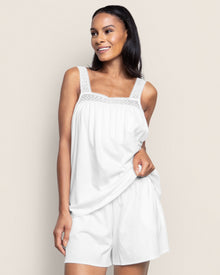 A woman wearing the white Womens Pima Camille Short Set by Petite Plume poses against a light background, smiling and gently holding her top adorned with lace straps and neckline.