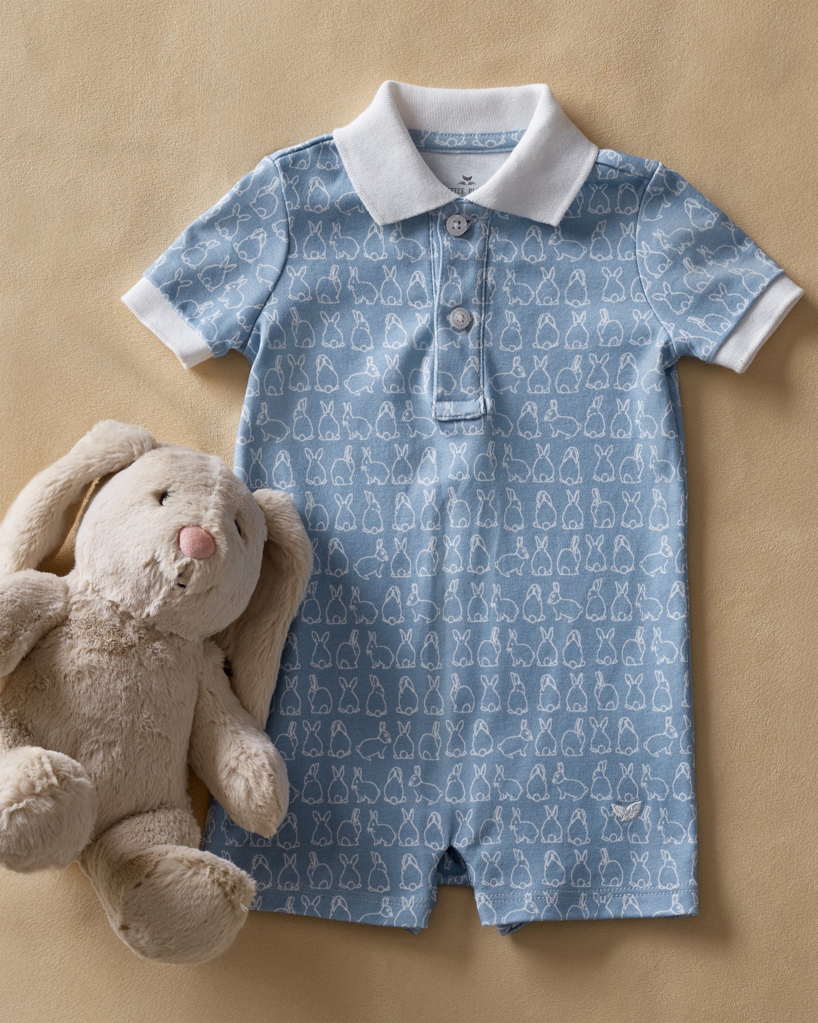 The Baby's Pima Louis Play Romper in Hoppy Days Blue is a light blue short-sleeve romper with a white collar and sleeve cuffs, a white bunny outline pattern, and bottom snaps for easy changing.
