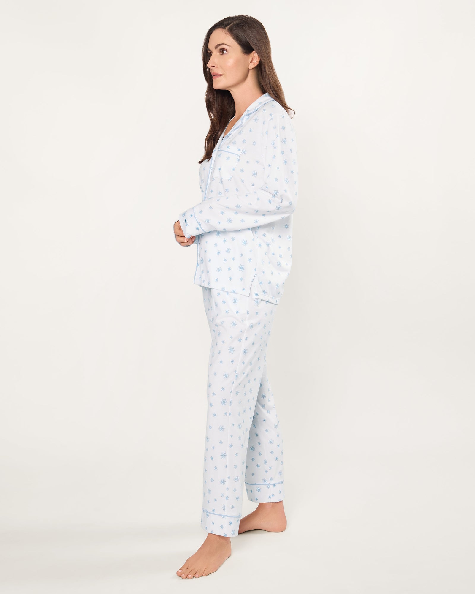The Women's Pima Pajama Set in Periwinkle Wonderland features white pajamas with light blue snowflake patterns, a collared long-sleeve button-up top with pocket, and matching elastic waist pants, both accented with blue piping.
