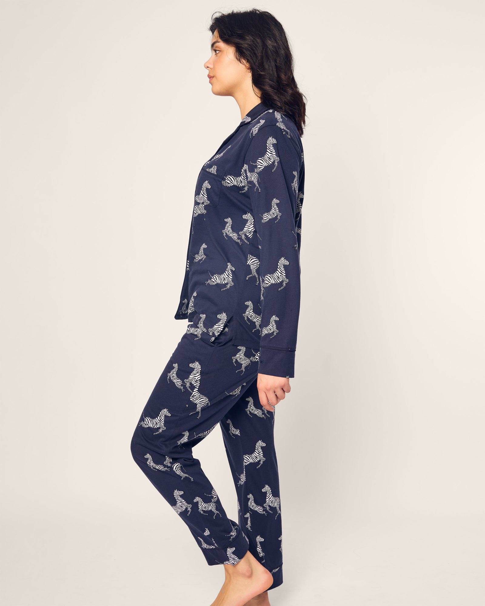 A person in profile stands barefoot against a plain backdrop, draped in the Womens Pima Pajama Set in Zebre de Nuit: navy blue luxury sleepwear adorned with elegant white zebra prints, crafted from soft Pima cotton.