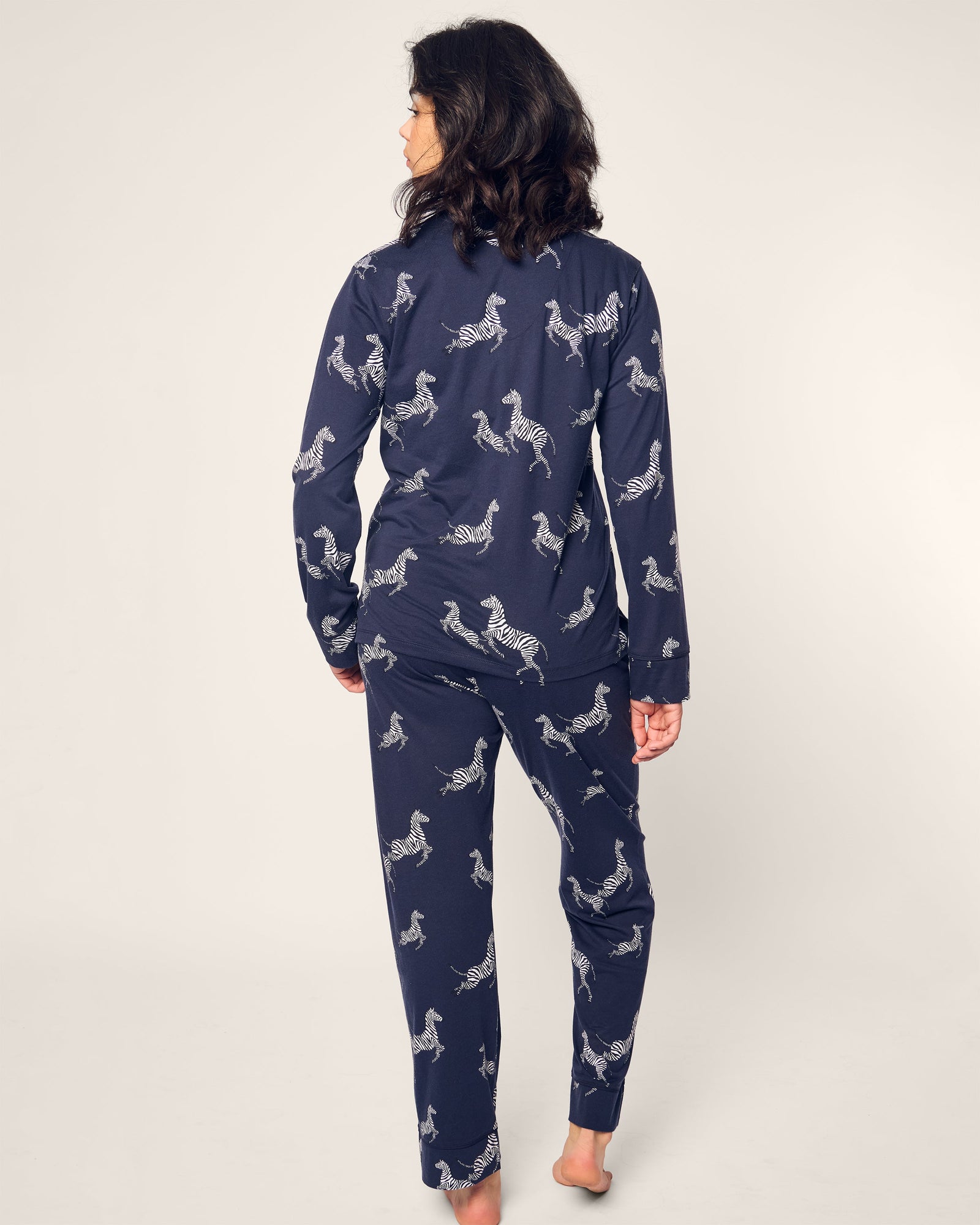 A person with dark hair stands barefoot, facing away, wrapped in a Womens Pima Pajama Set in Zebre de Nuit by Petite Plume, featuring luxury navy-blue zebra print on an off-white background.