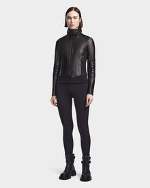 A woman stands facing forward wearing the Rudsak PRESLEY - 8122027 BLACK leather jacket, crafted from supple lamb leather, paired with black leggings and black ankle boots against a light background.