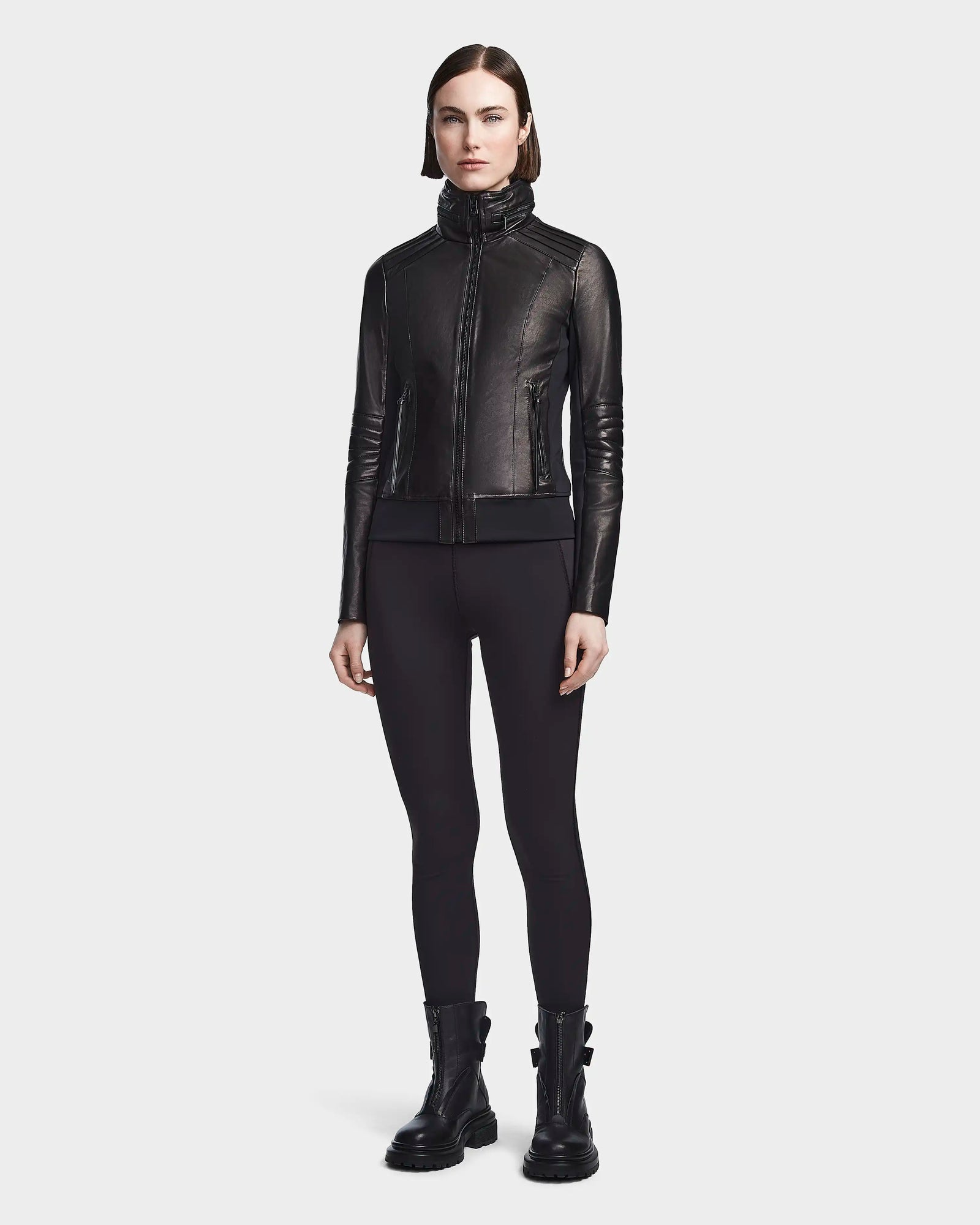 A woman stands facing forward wearing the Rudsak PRESLEY - 8122027 BLACK leather jacket, crafted from supple lamb leather, paired with black leggings and black ankle boots against a light background.