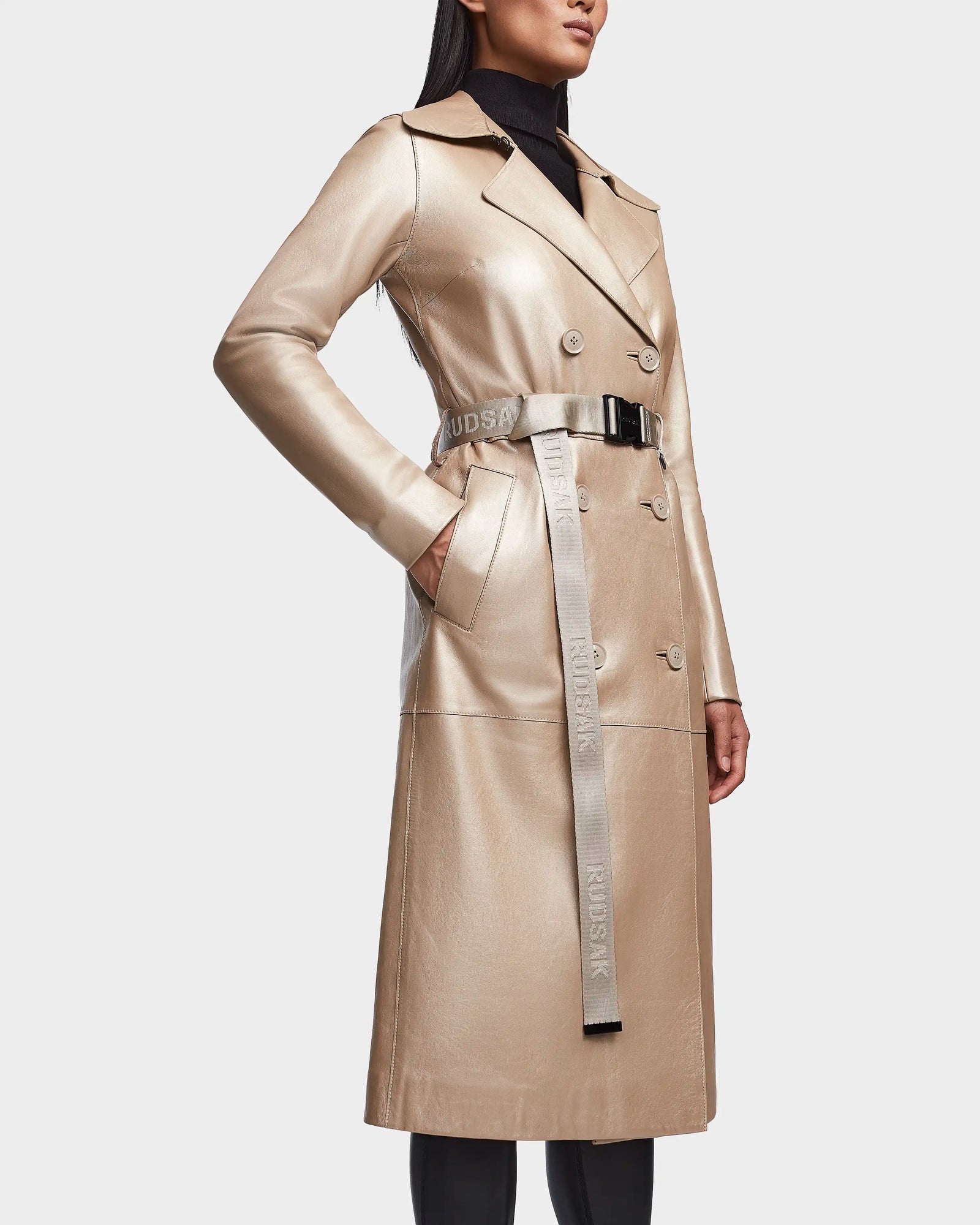 A woman wears the Rudsak KIRALY - 8123553 Metallic Ivory leather trench with a black turtleneck. The coat features a double-breasted design, belted waist with RUDSAK text, and side pockets for a chic, modern look.