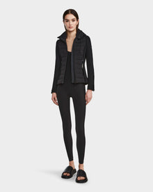 A woman faces forward against a plain white background wearing the RUDSAK MABS - 8122553 BLACK women’s puffer jacket, styled with a black long-sleeve top, leggings, and slide sandals.