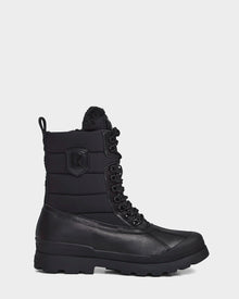 The REAGAN - 6222500 BLACK by Rudsak is a black mens mid-calf winter boot with a rugged rubber sole, padded upper, side patch detail, and faux fur lining at the top—crafted for warmth and style against a white background.