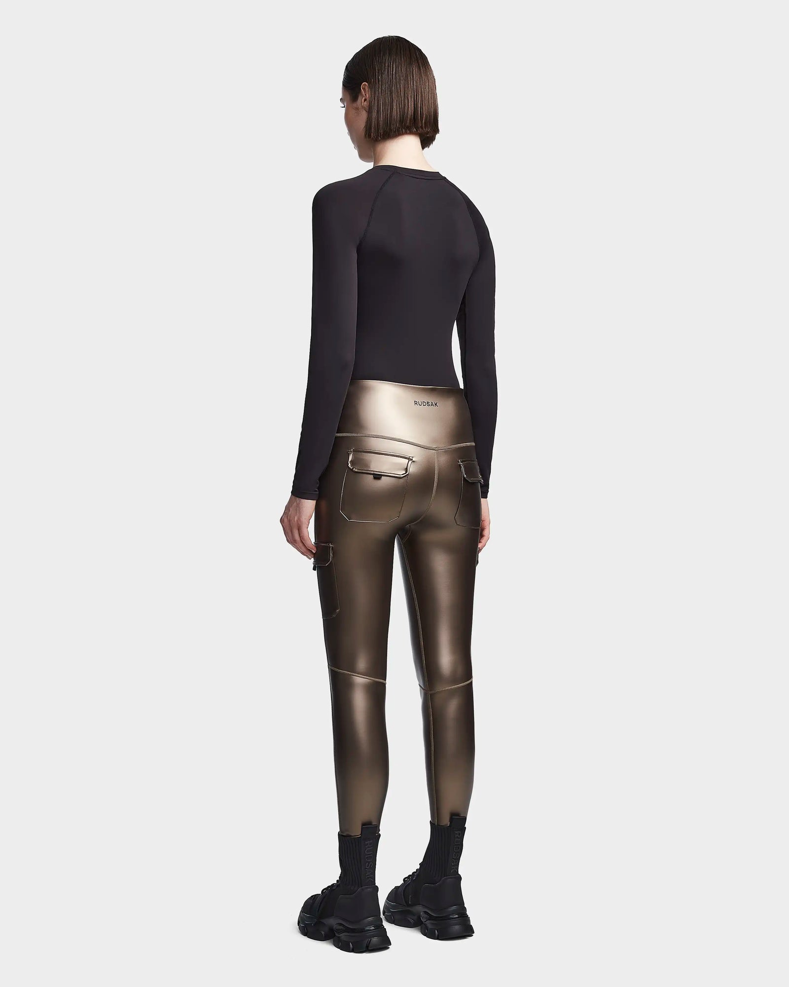 A person with short brown hair faces away, wearing a black long-sleeve top, Rudsak SIEN P - 8523027 METALLIC CHOCOLATE stretch vegan leather cargo leggings with pockets, and black chunky sneakers against a plain light background.