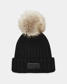 The ANISA - L423500 Black/Natural by Rudsak is a black knit beanie with a wide ribbed pattern, folded brim, and large faux fur pom pom. Finished with a rectangular black Rudsak patch on the front, it’s perfect for winter style.