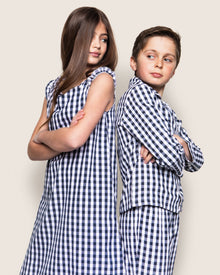 A girl with long hair and a boy with short hair stand back-to-back with arms crossed, wearing matching outfits. The girl is in Petite Plumes Girls Twill Amelie Nightgown in Navy Gingham, and both look over their shoulders playfully.