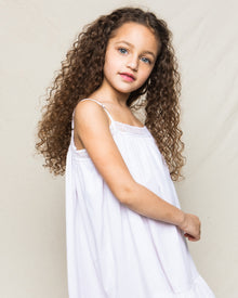 A young girl with long curly hair, wearing a Petite Plume Girls Twill Lily Nightgown in White, stands against a plain light background, slightly smiling at the camera.
