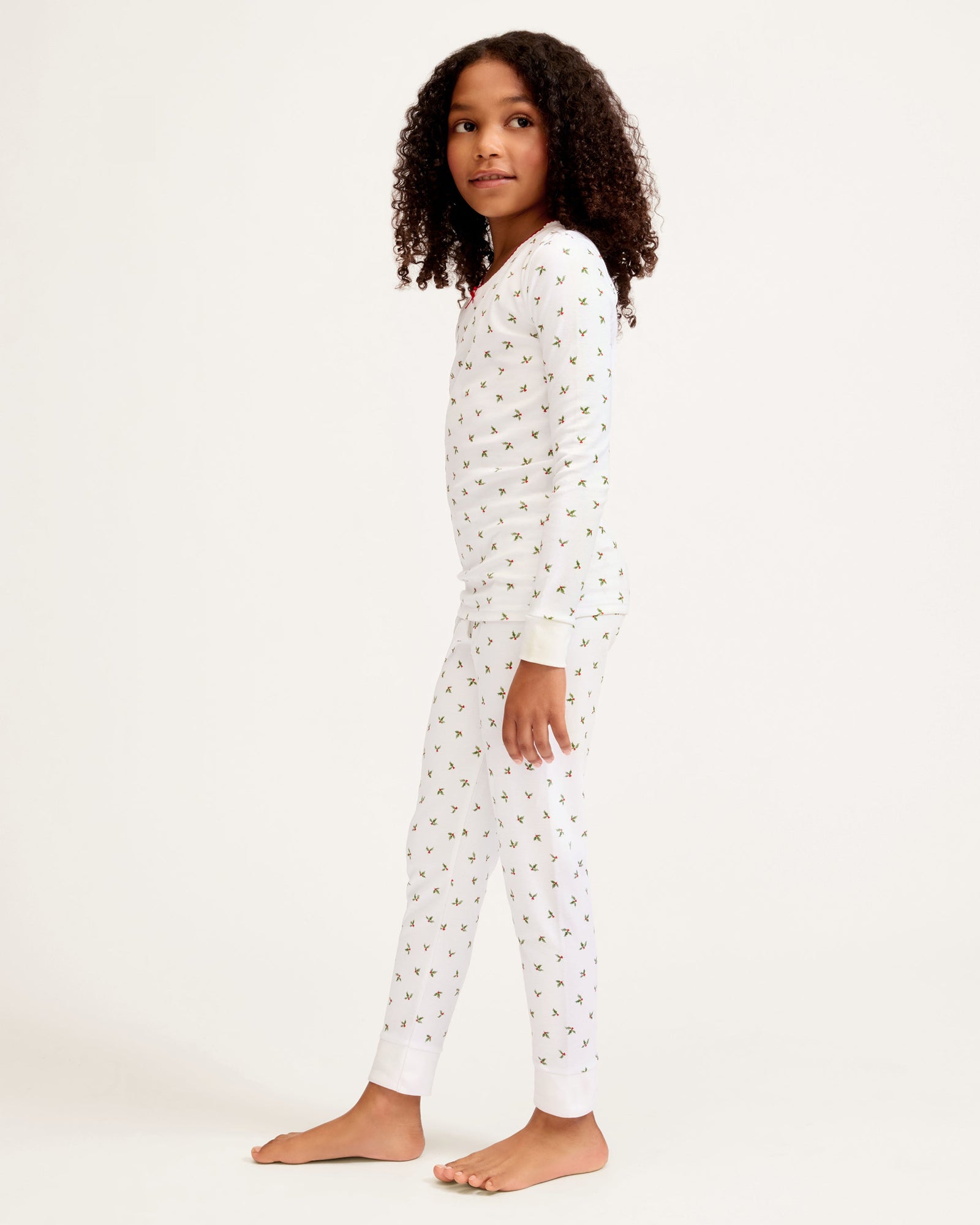 Kid's Pima Snug Fit Pajama Set in Mistletoe Moments: a two-piece white pajama set for children with a long-sleeve top, pants, green and red holly pattern, red scalloped neckline, and a red bow at the collar.