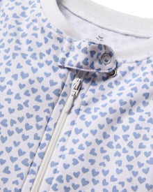 Close-up of Petite Plumes Babys Pima Romper in Bluehearts, highlighting a snug fit with blue hearts on white. Made from soft Pima cotton, it has a white front zipper and a snap button closure at the top.