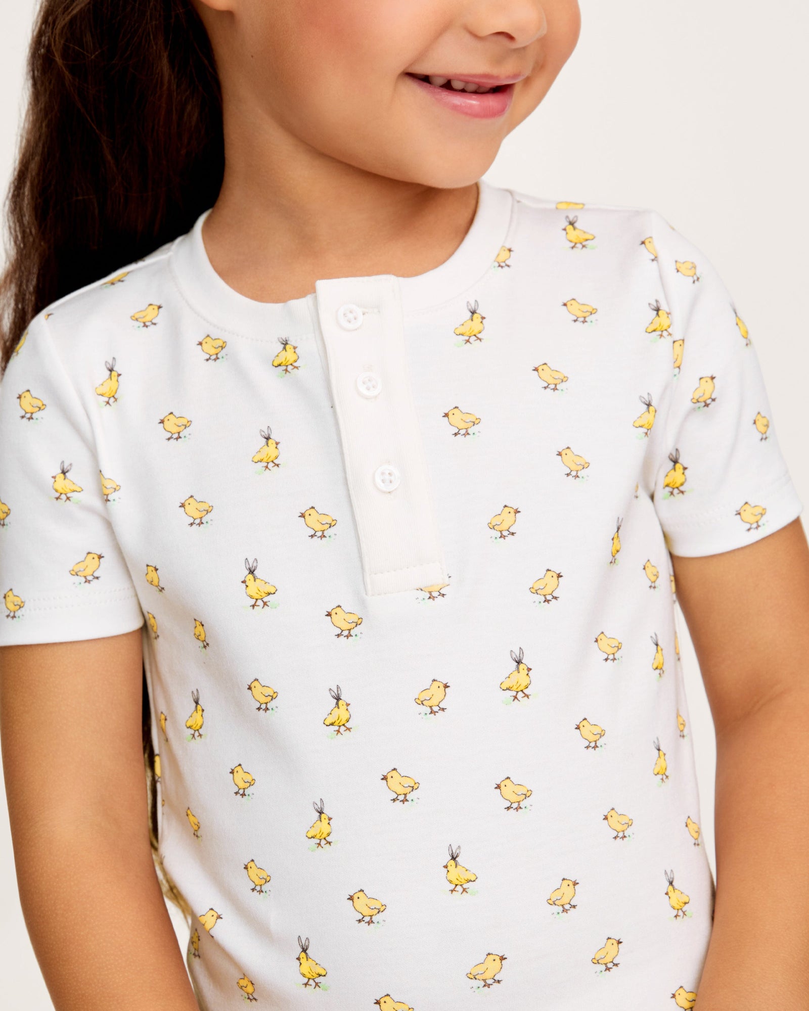 A young child with long brown hair smiles while wearing the Kid's Pima Snug Fit Short Set in Tres Chick, standing against a plain light background.