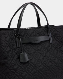 Close-up of the Rudsak ASMINE - L325005 Black tote with an embossed monogram pattern, two leather handles, a leather tag, and a branded leather accent near the top.