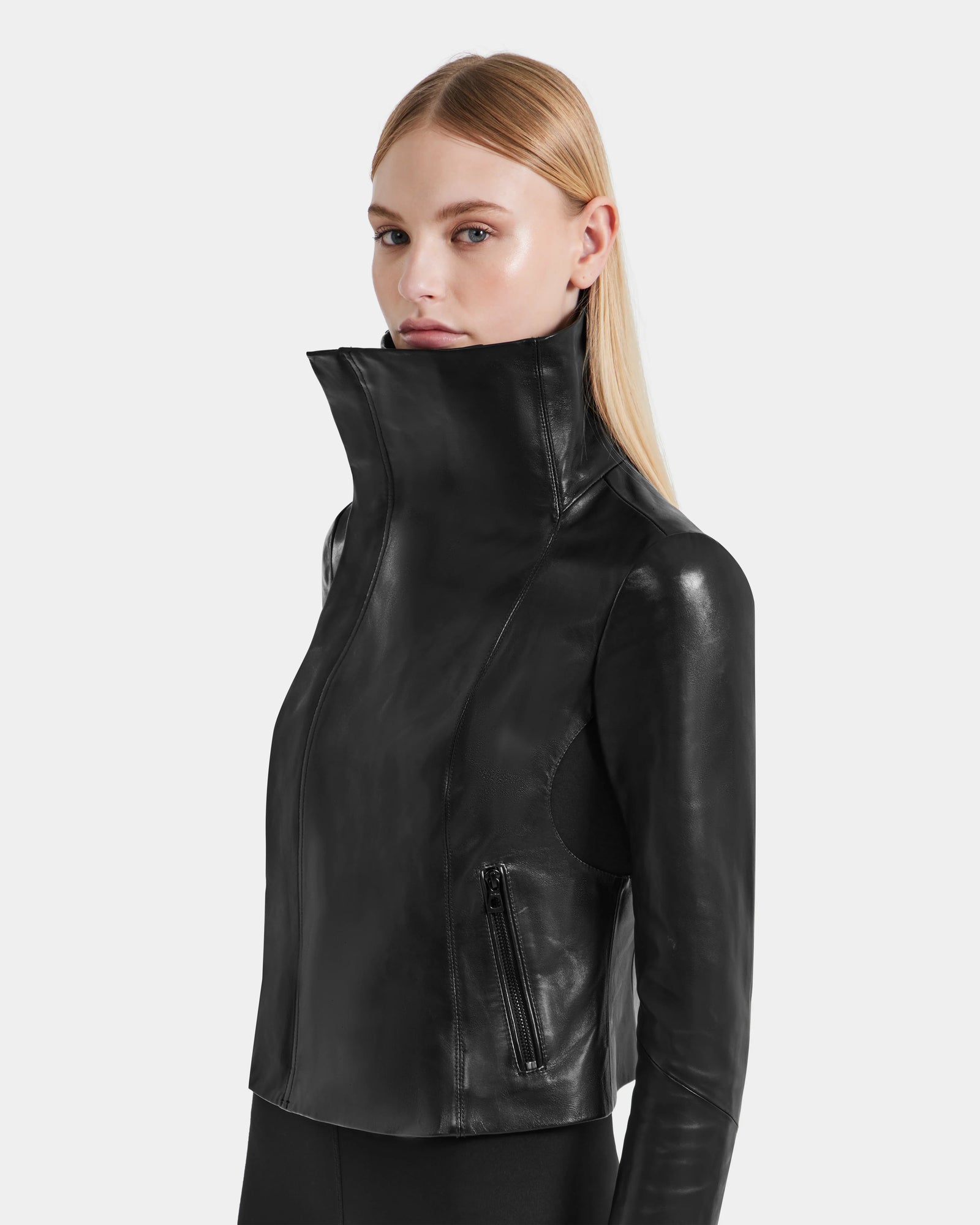 A woman with straight blonde hair wears the Rudsak AVRIL - 8124561 Black leather jacket with a high collar and zippered pockets, standing against a plain light gray background.