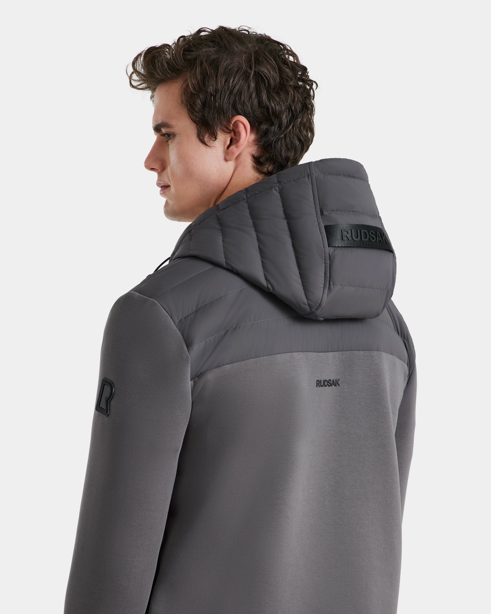 A man with short brown hair models the Rudsak DEE MAT - 6124028 Grey men’s lightweight down jacket, featuring a quilted hood and visible back logo, shown from behind at a slight angle against a plain white background.