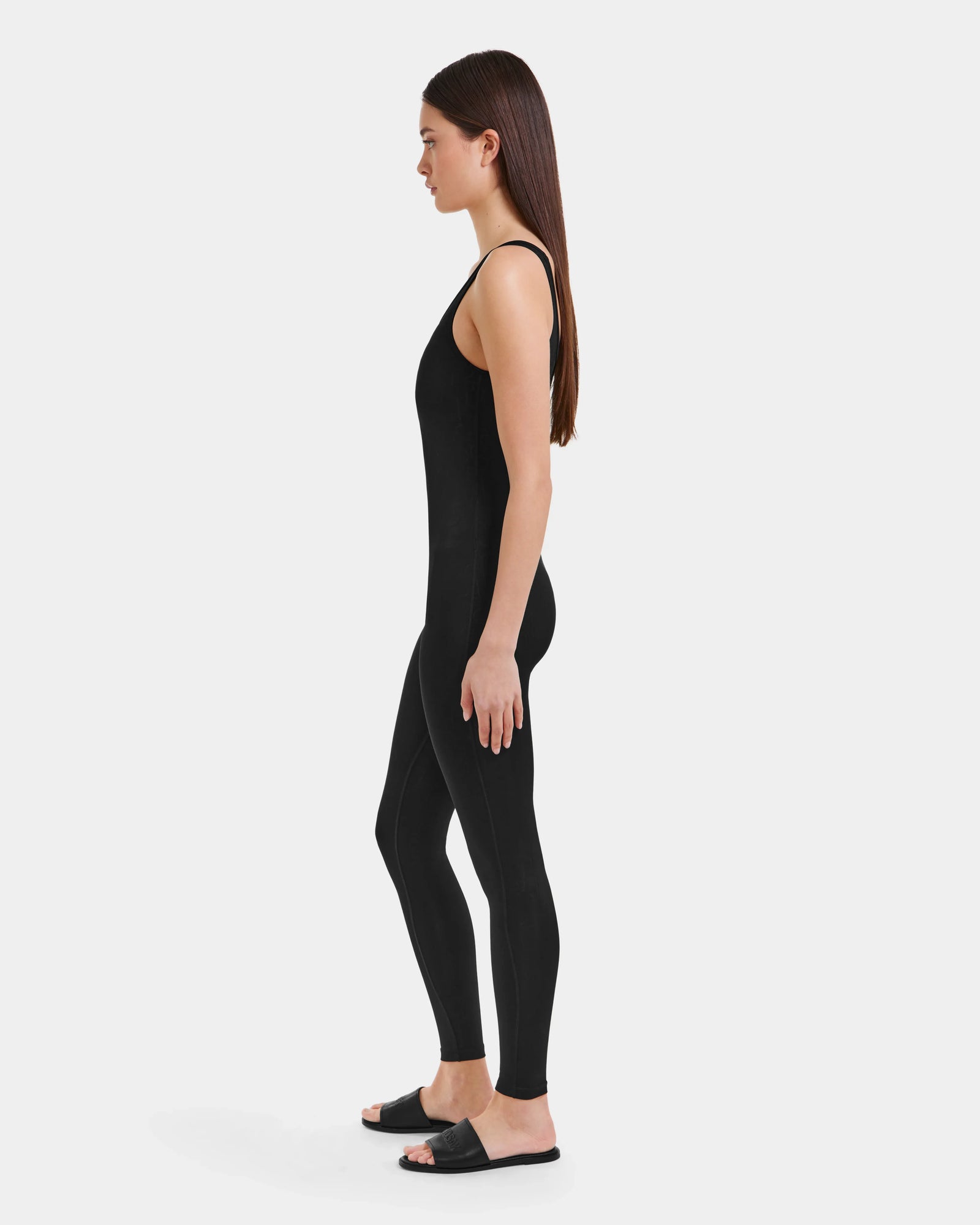 A woman in profile against a white background wears the LAWREN - 8523054 Rudsak Pattern bodysuit by Rudsak, styled as a jumpsuit, with black slide sandals.