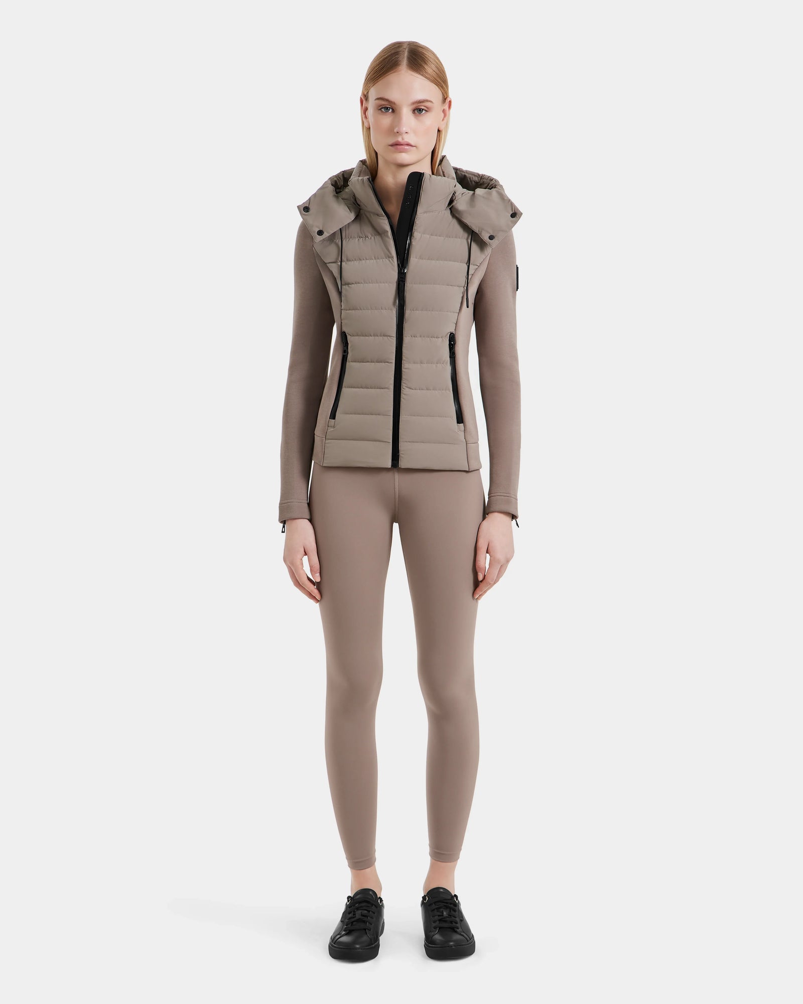 A woman faces forward, wearing Rudsaks MABS - 8124825 Cement taupe puffer vest over a lightweight jacket, paired with a long-sleeve taupe top, matching fitted pants, and black sneakers against a white background.