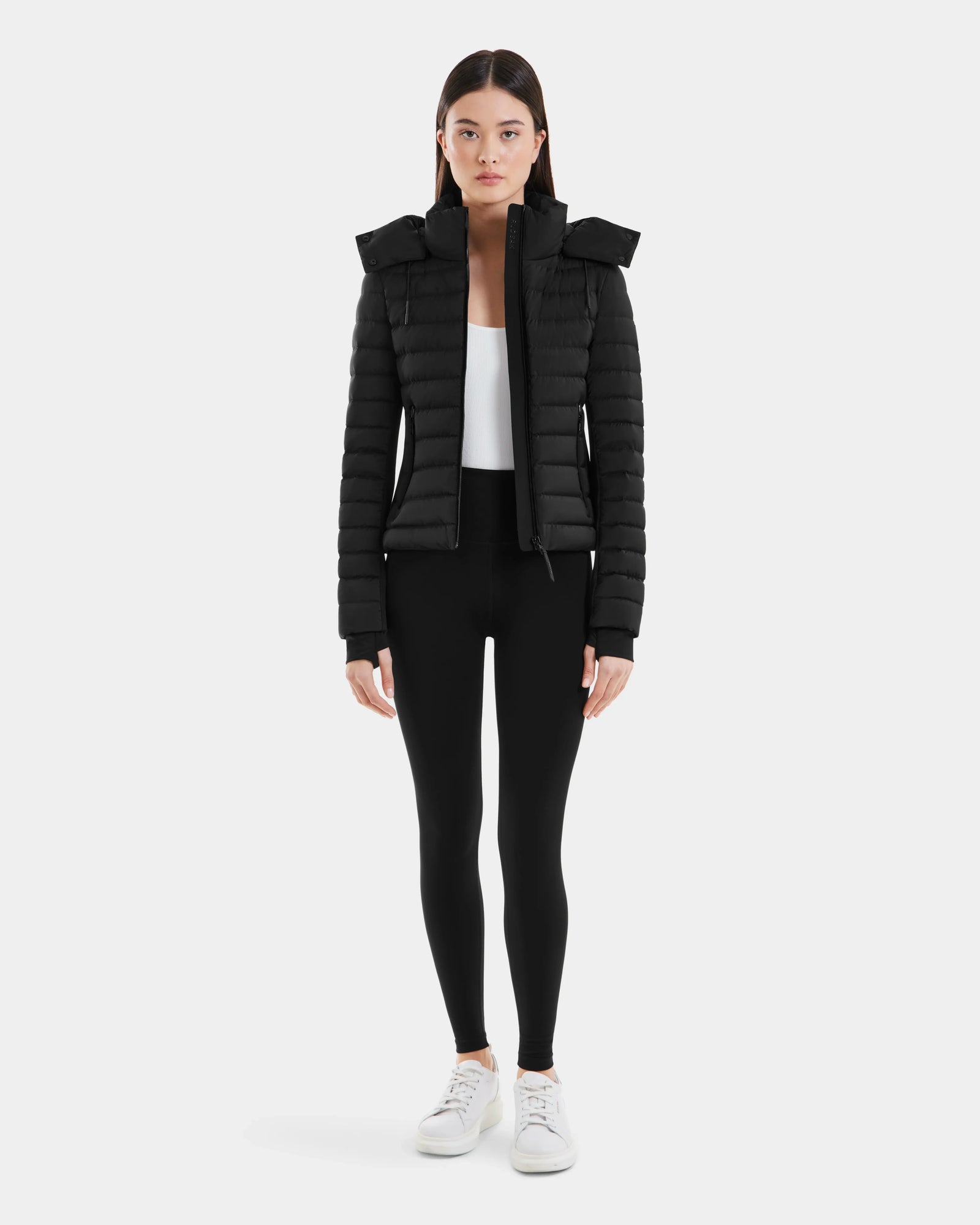 A woman faces forward wearing the Rudsak MARYA M - L123559 Black down puffer jacket, paired with black leggings, a white top, and white sneakers against a plain light background.