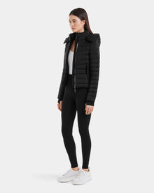 A woman stands slightly to the side against a plain white background, wearing the Rudsak MARYA M - L123559 Black women’s down puffer jacket, black leggings, a white top, and white sneakers.
