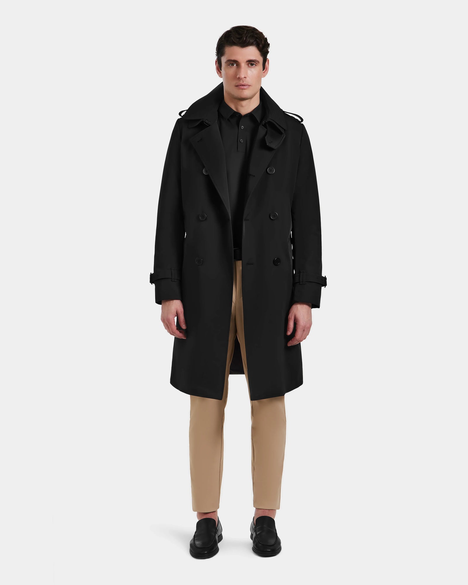 A man stands facing forward against a plain white background, wearing the Rudsak TIMOTHY - 6124589 Black mens trench coat over a black shirt, tan pants, and black shoes.