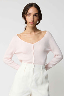 Woman wearing a light pink long-sleeve top and white pants on a plain background