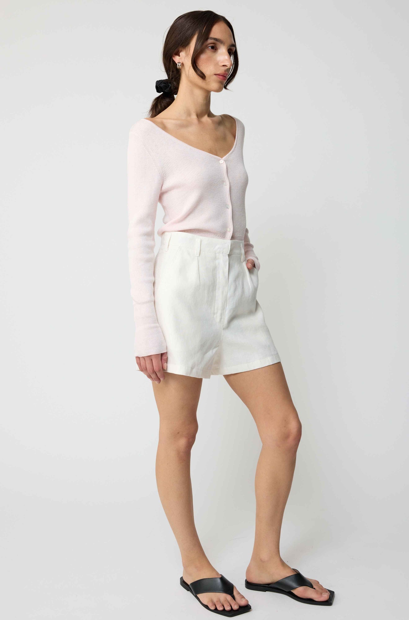 Woman wearing a light pink cardigan and white shorts on a plain background