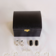 Black jewelry box with gold clasp, black earrings, and silver rings on a white background