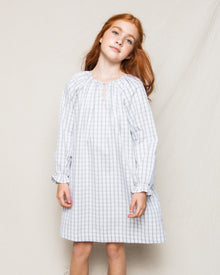 A young girl with long red hair stands against a plain backdrop in Petite Plumes Girls Twill Delphine Nightgown in Regent Tattersall, reminiscent of luxurious sleepwear. She tilts her head slightly, gazing forward with serene confidence.