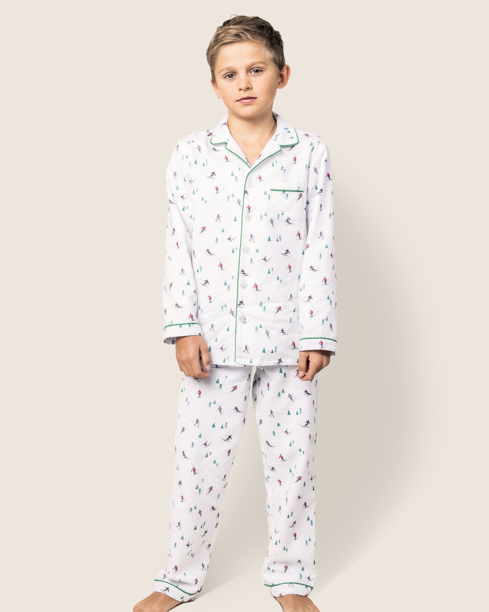 A young boy stands barefoot against a plain background, wearing Petite Plumes cozy Kids Flannel Pajama Set in Apres Ski. These flame-retardant pajamas feature colorful designs, a classic collar, and front buttons for a charming look.