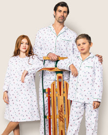 A man, a young girl, and a boy wear matching Petite Plumes Kids Flannel Pajama Set in Apres Ski print, safe with flame-retardant fabric. The man and boy rest their hands on a wooden sled against a neutral background.