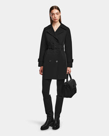 A woman wearing the Rudsak KIMBERLY - 8124801 Black belted trench coat, black leggings, black knee-high boots, and holding a black handbag stands against a plain light background.