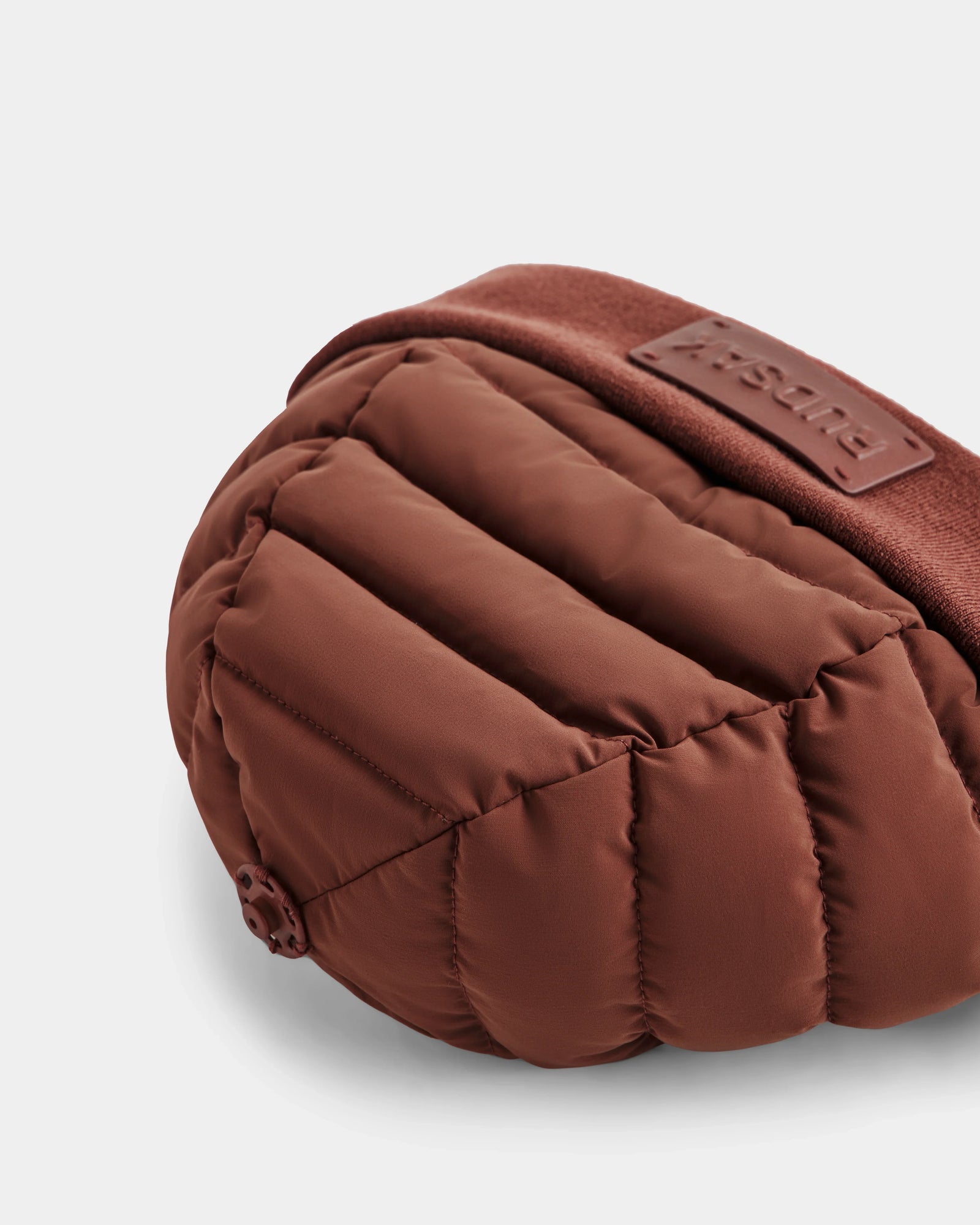 A close-up of the Rudsak GALA - 8424514 Rust bag shows its quilted, padded brown fabric with horizontal stitching and a stitched logo patch on the matching strap, set against a plain white background.