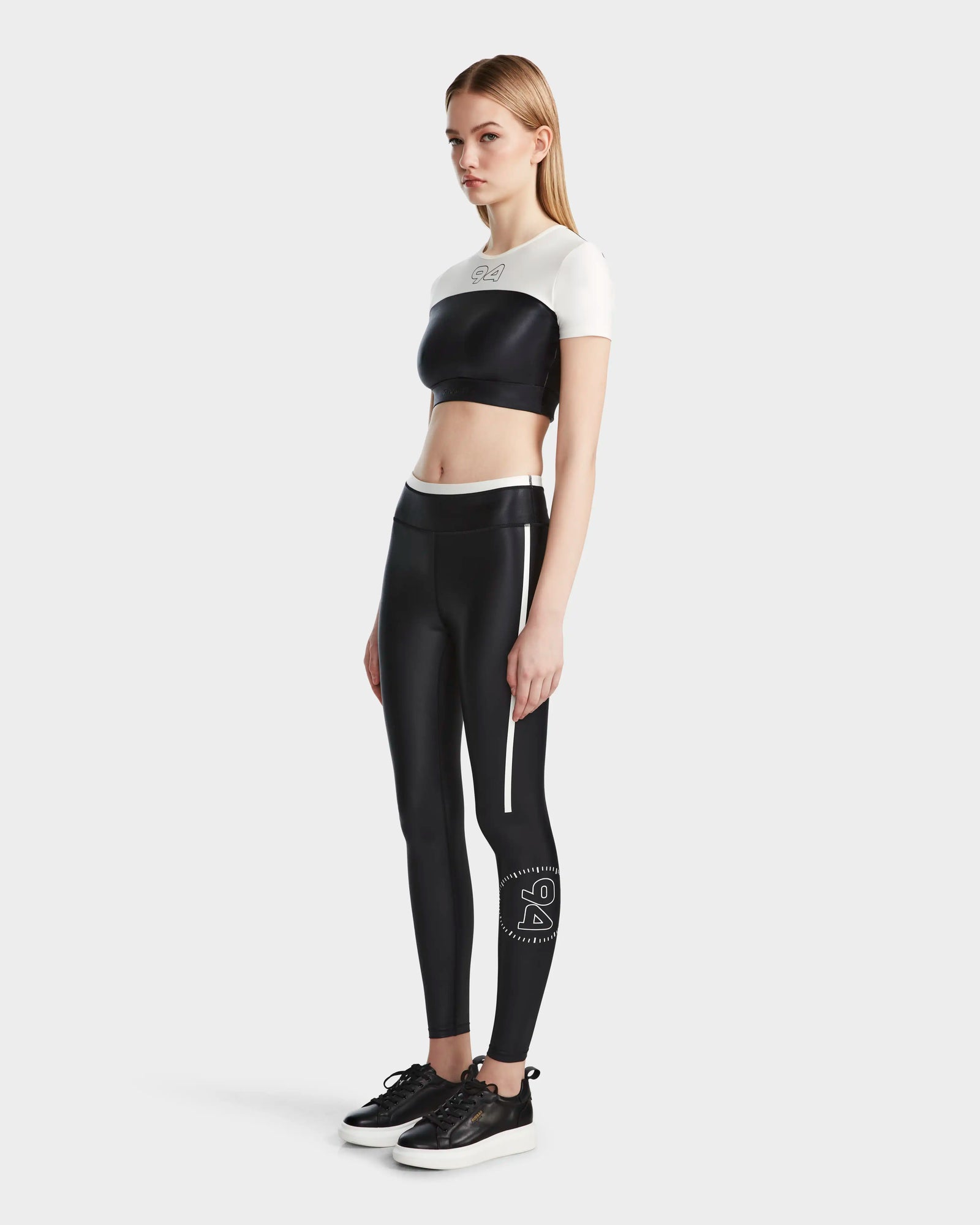 A woman wears the Rudsak LILIANE - 8524018 Black workout crop top and high-waisted black leggings with a logo on the left leg, paired with black sneakers, posing confidently against a plain light gray background.
