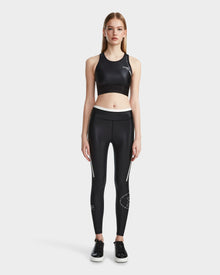 A woman stands facing forward wearing Rudsaks TRACY - 8524016 Black high-performance sports bra styled as a black crop top, paired with matching black high-waisted leggings with white details and black sneakers, set against a plain white background.